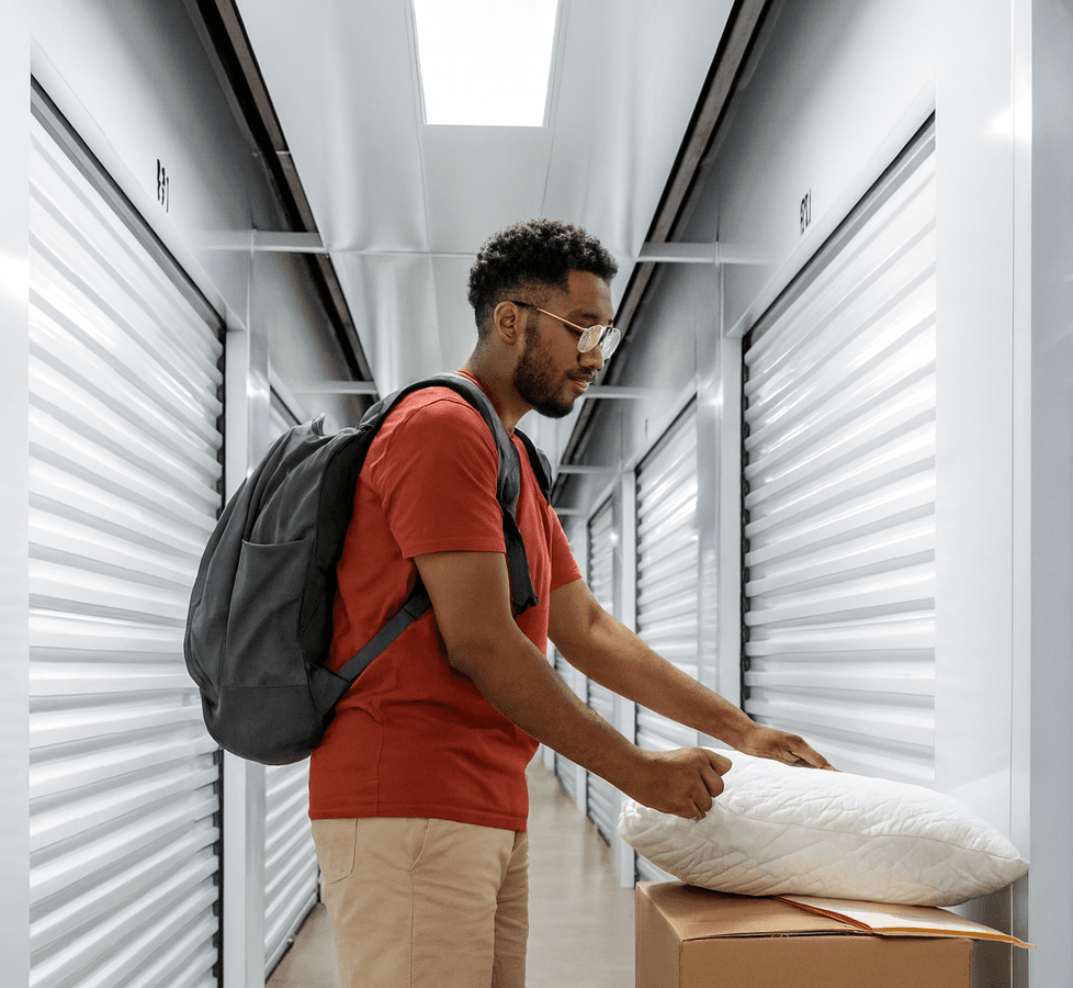 A college student with a backpack places boxes and a pillow outside a storage unit in a clean, well-lit indoor facility.